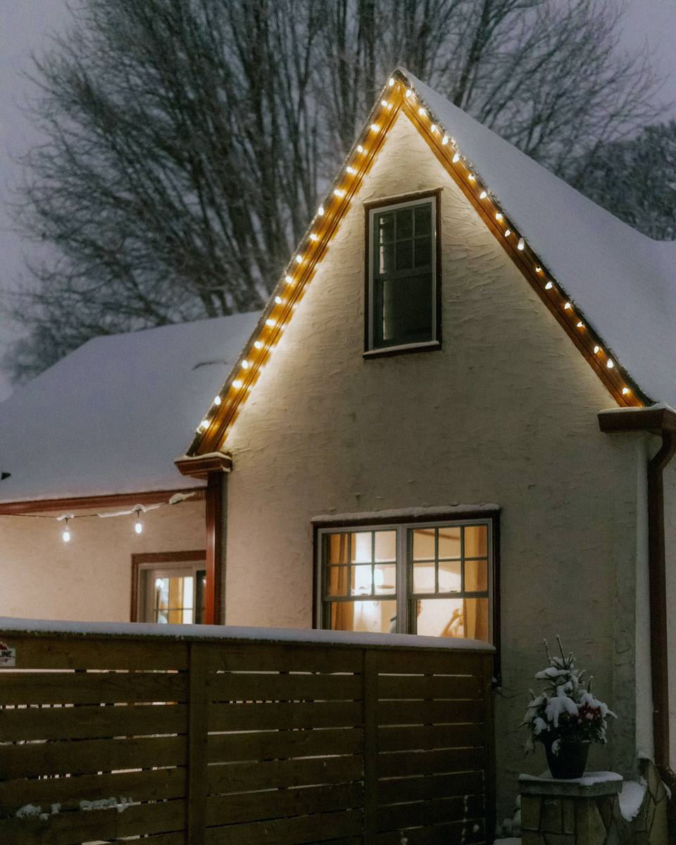 Snow-covered home at dusk, its roofline traced in warm white C9 bulbs, windows glowing from inside